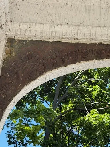 The carved acanthus band along the porch arch stripped to dark bare wood, with trees beyond.